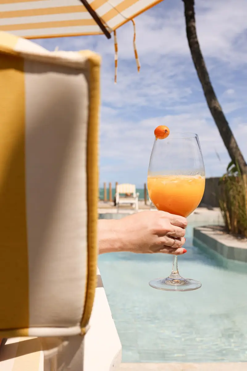 Mão segurando um drink laranja na borda da piscina